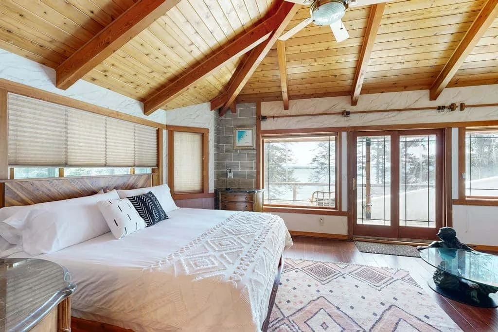 A bedroom with a bed beneath a sloped wooden ceiling and large windows.