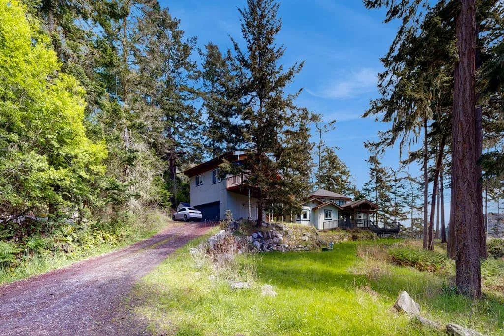A house sits beside a gravel driveway and grassy lawn among tall trees.