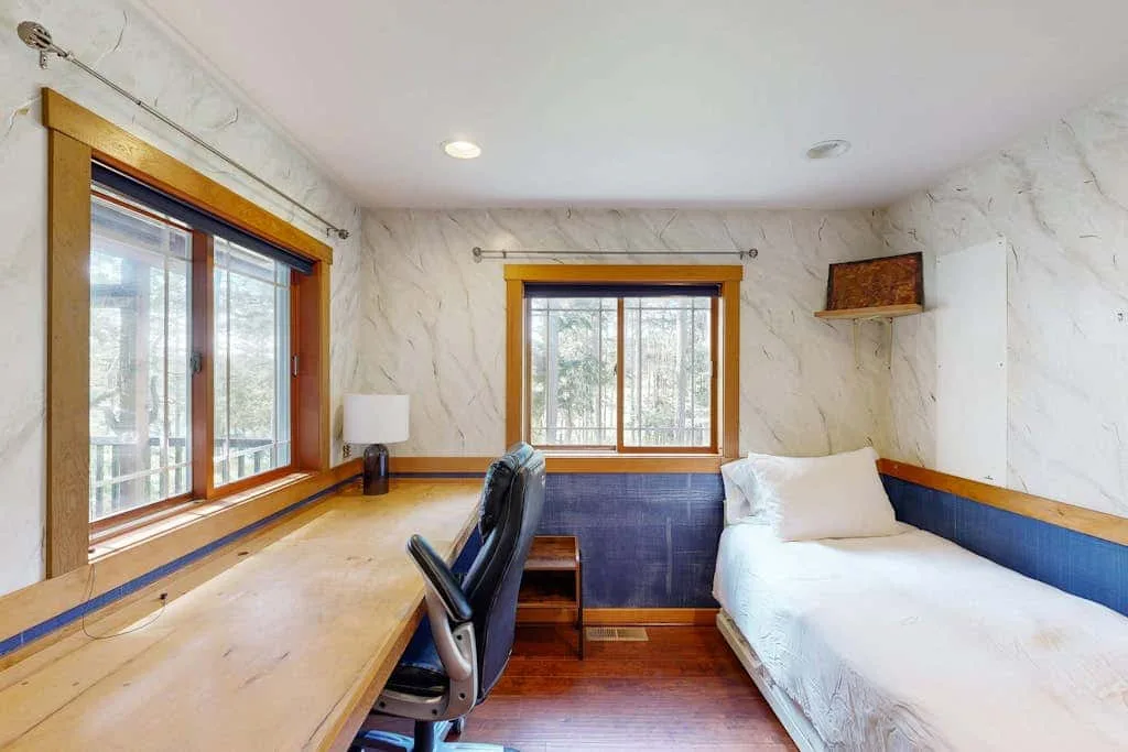 A compact bedroom workspace has a desk beside a bed and two windows.