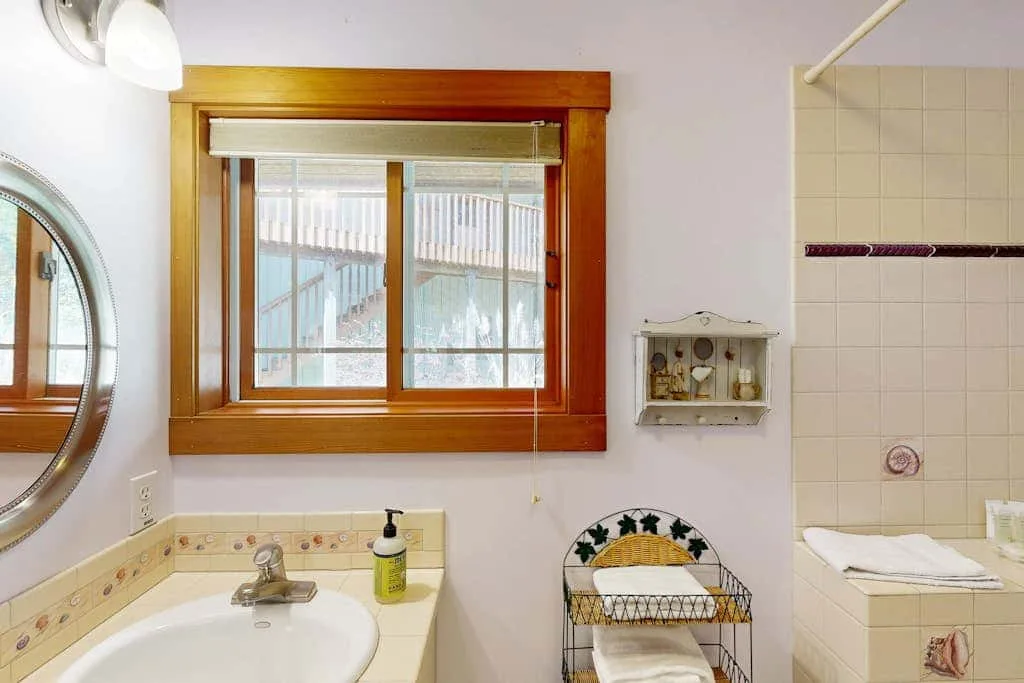 A bathroom vanity sits beneath a wood-trimmed window, with a tiled bathtub nearby.