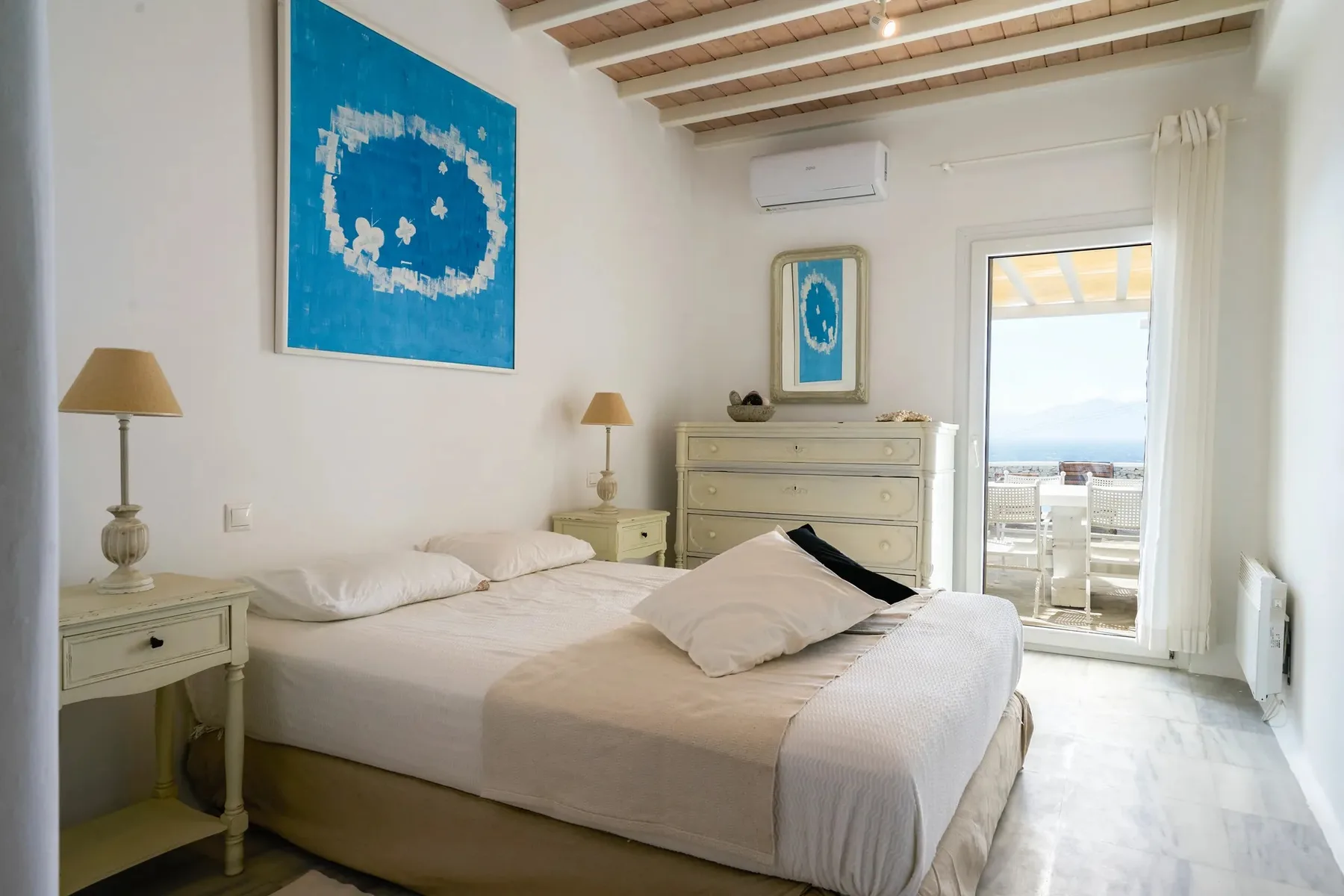 A bright bedroom with a bed, pale furnishings, and artwork on the walls.