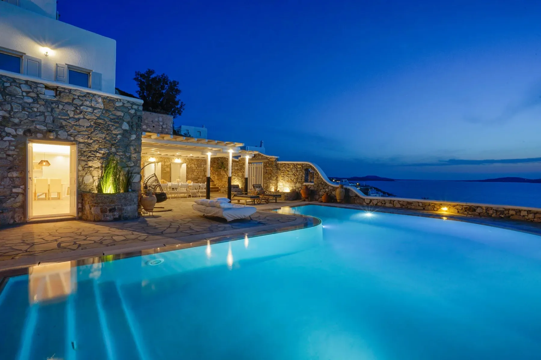 An illuminated outdoor pool beside a stone terrace at dusk.