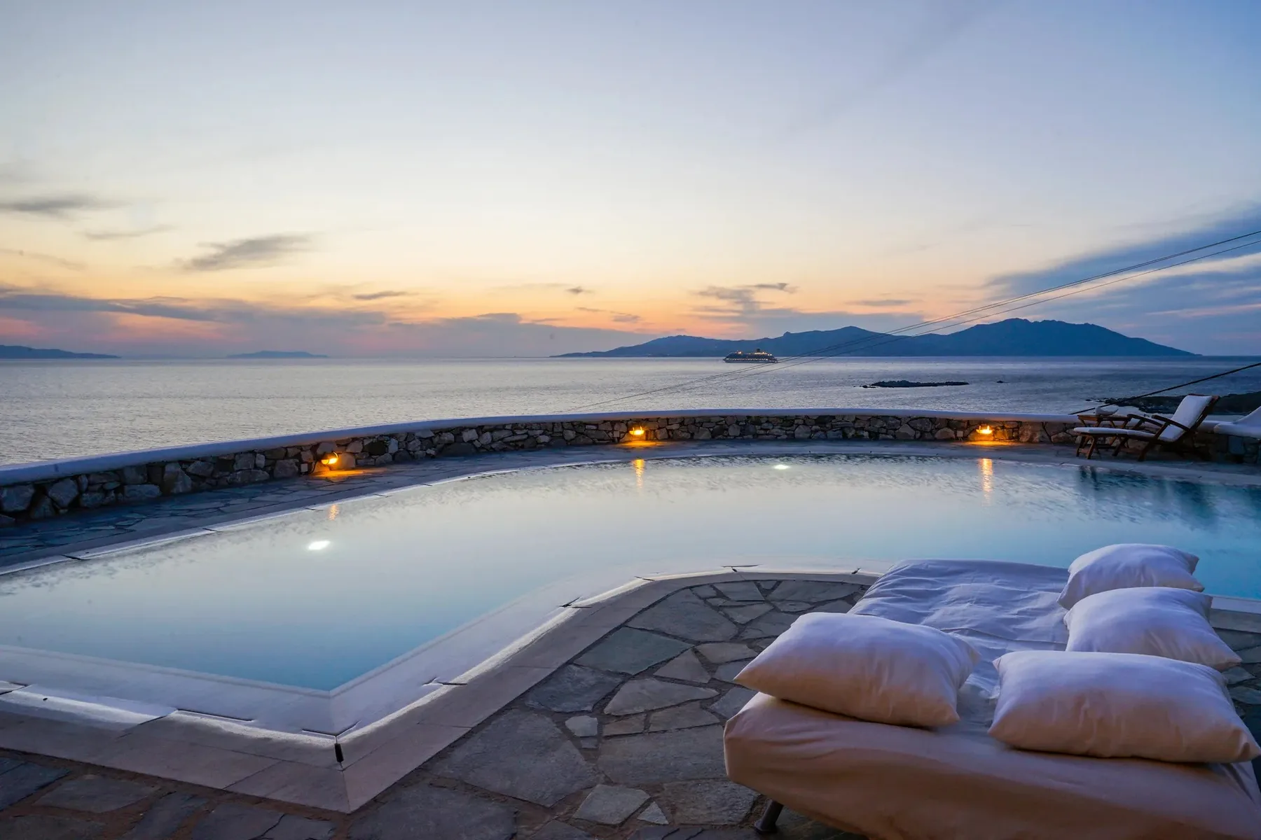 An outdoor pool overlooks the sea at sunset, with cushioned seating beside it.