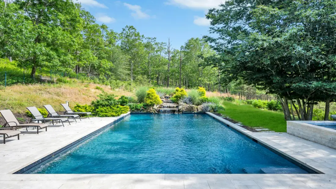 An outdoor swimming pool sits beside a paved patio and landscaped greenery.