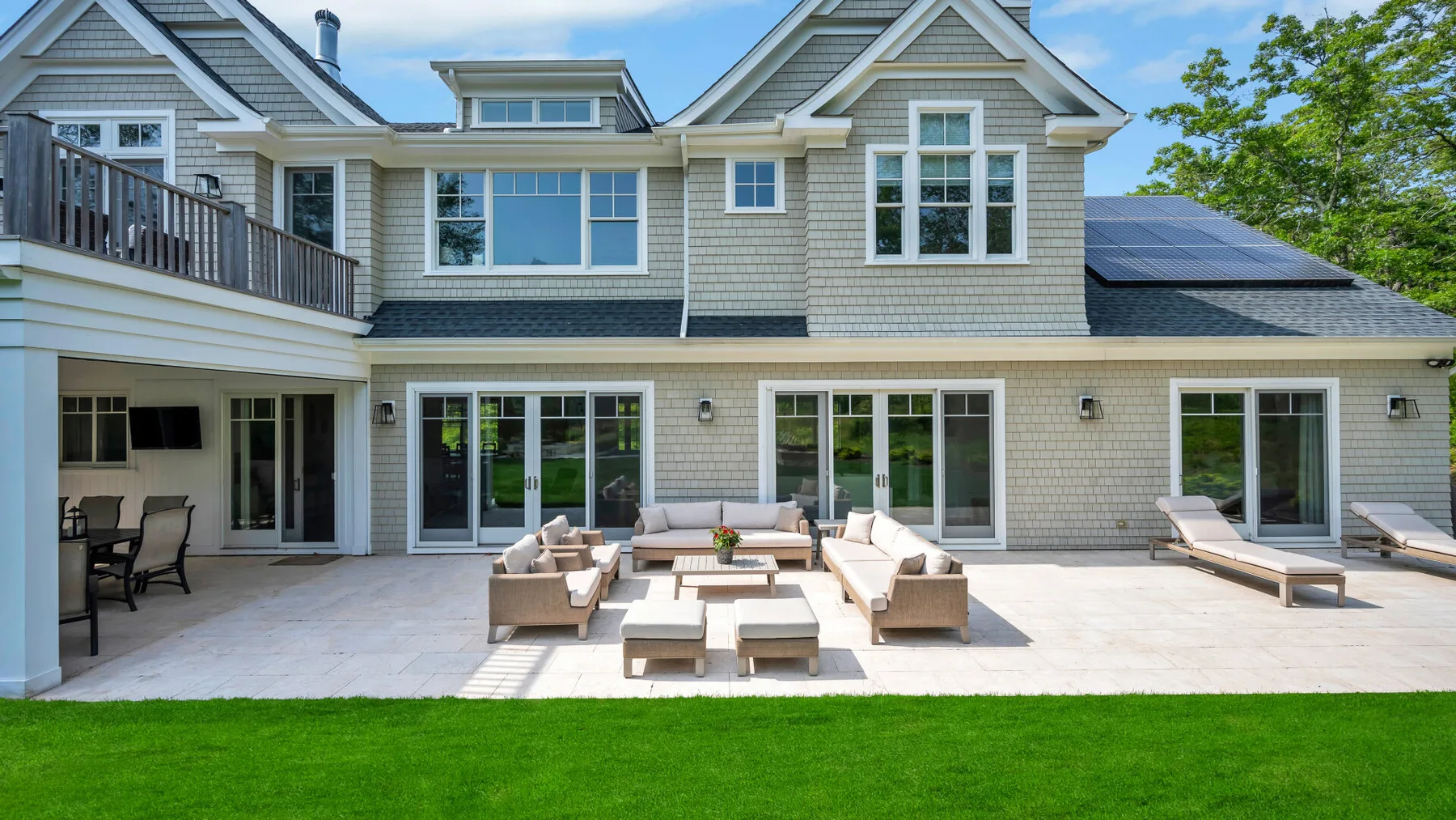 A large house opens onto a paved patio with outdoor seating and a green lawn.