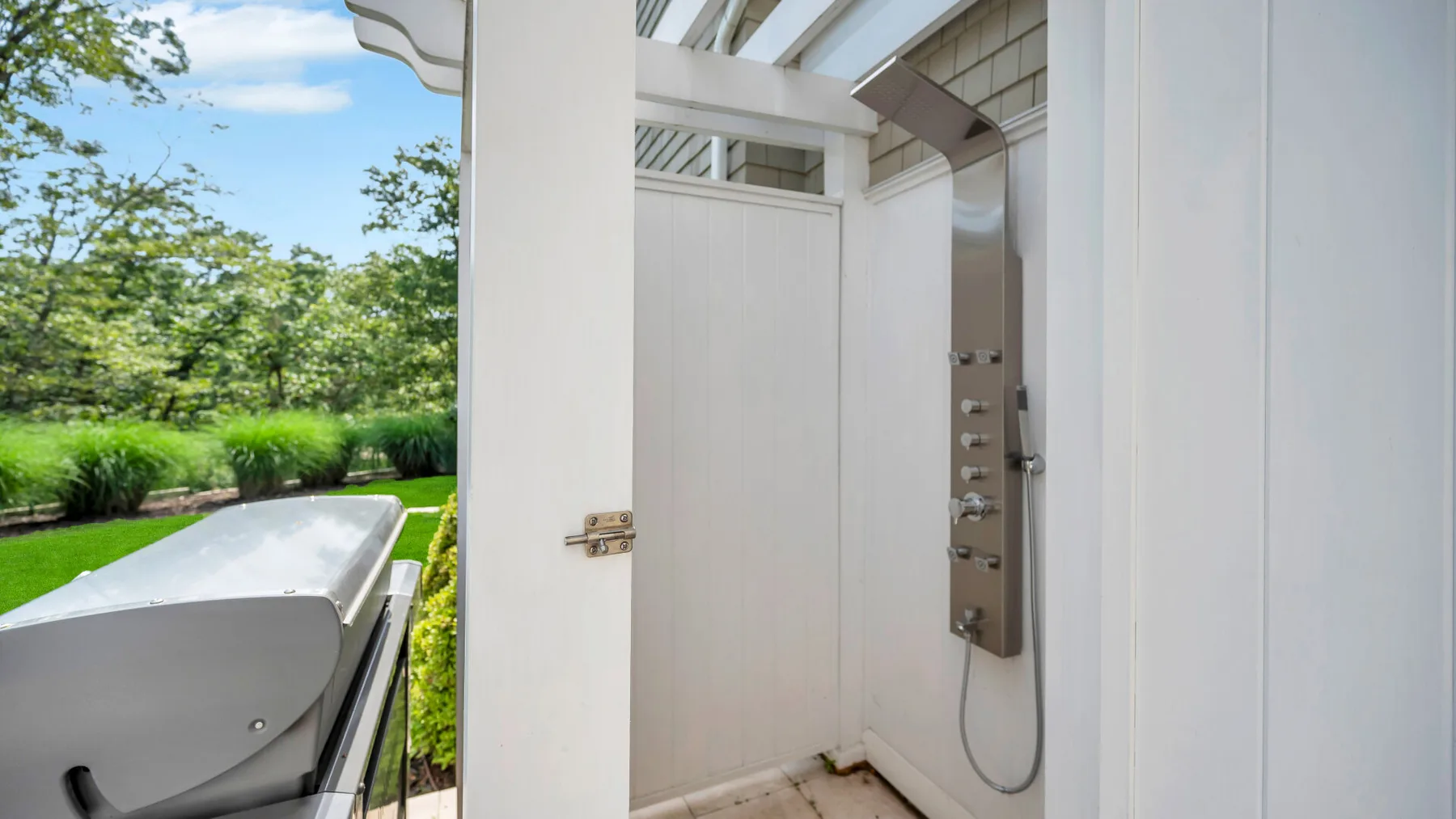 An outdoor shower stands beside a garden, with a metal control panel.