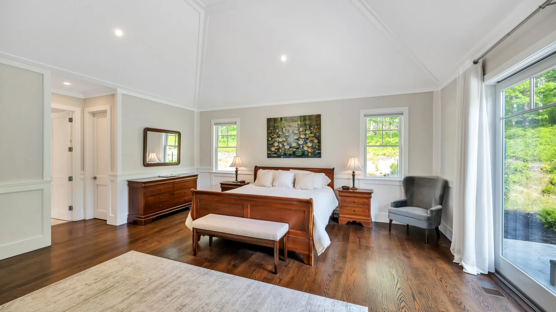A bright bedroom with a wooden bed, bench, and windows overlooking greenery.
