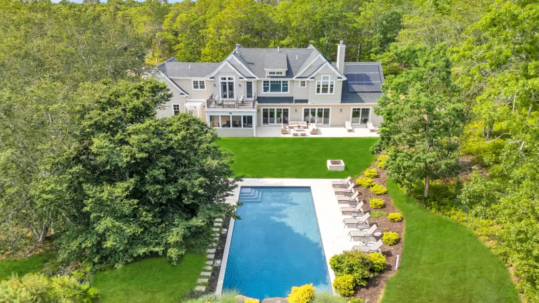 Una casa grande da a una piscina y un césped verde rodeados de árboles.