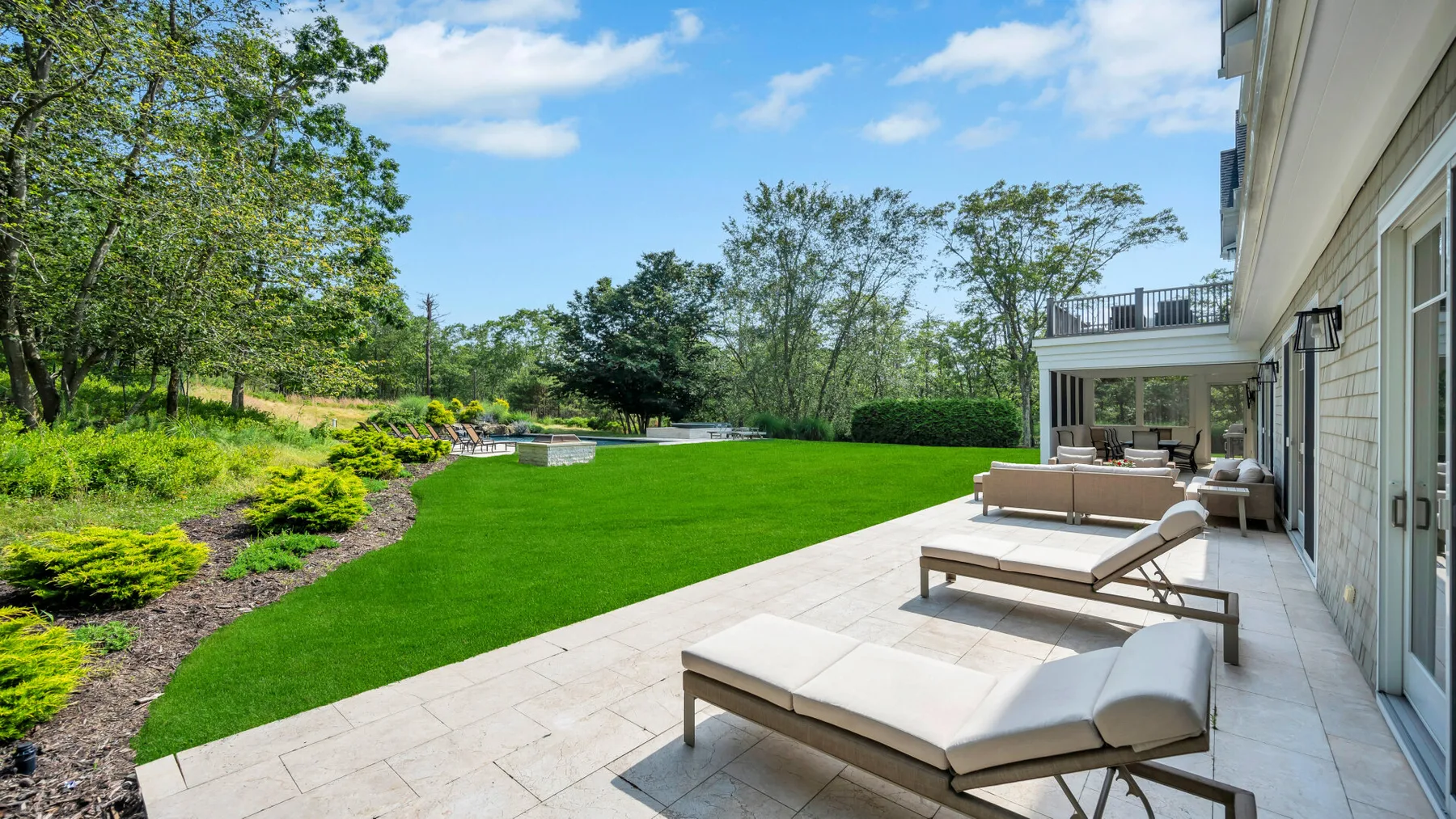 A landscaped backyard lawn borders a stone patio with outdoor seating.