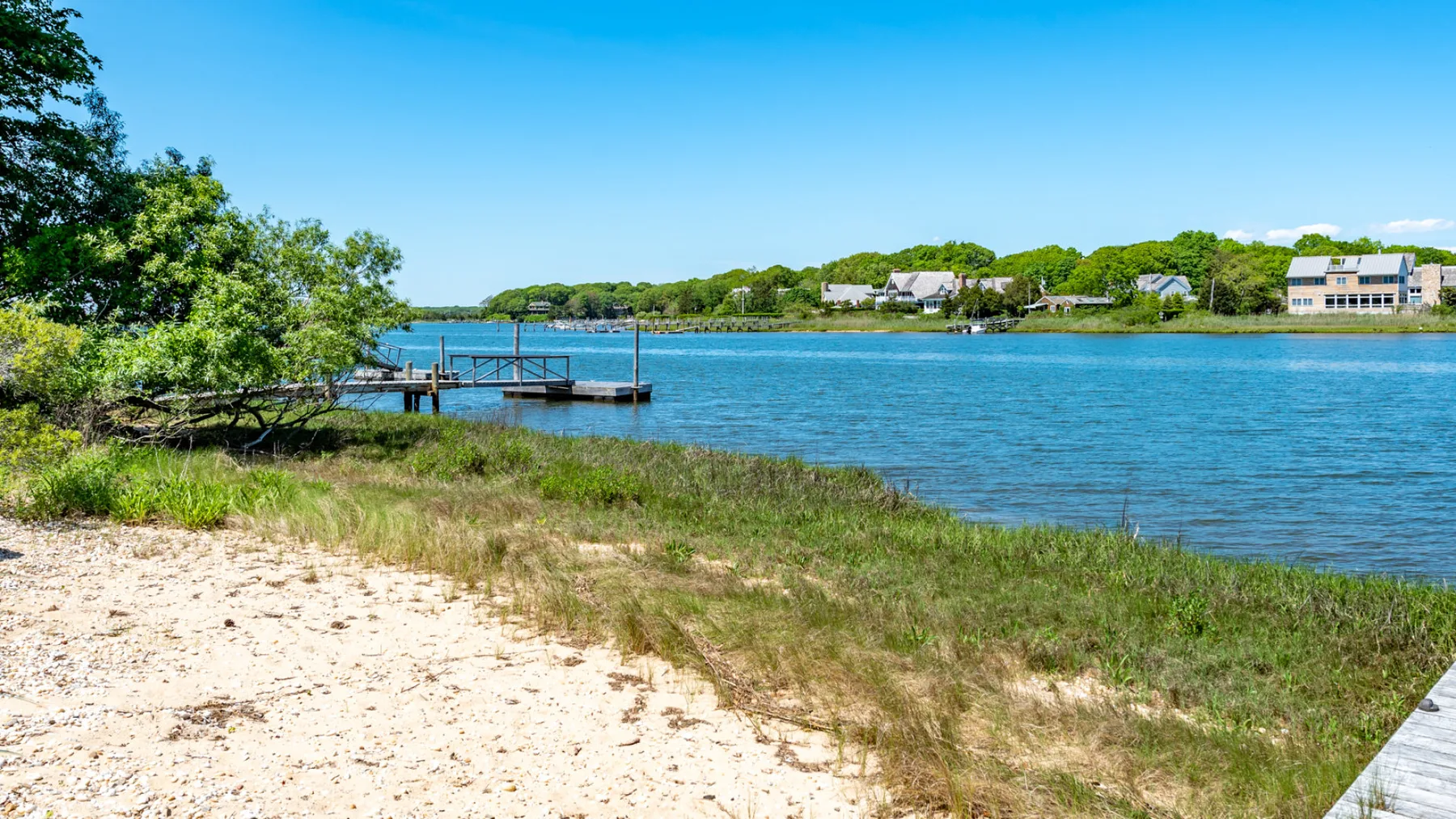 A sandy shoreline borders blue water, with a small dock and trees nearby.