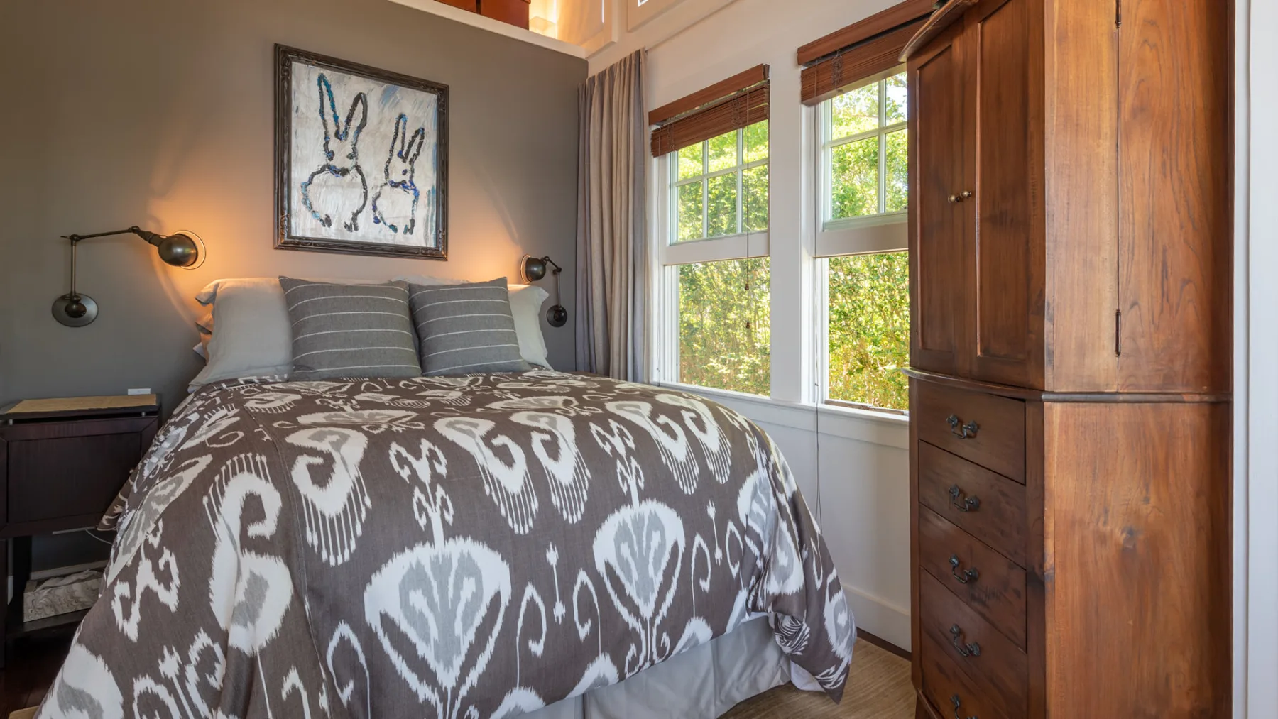 A bedroom with a bed beneath framed artwork and beside two bright windows.