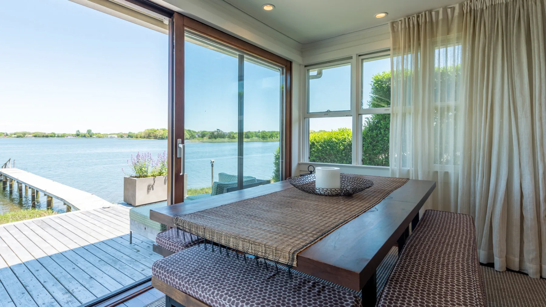 A dining area with a long table beside large windows overlooking the water.