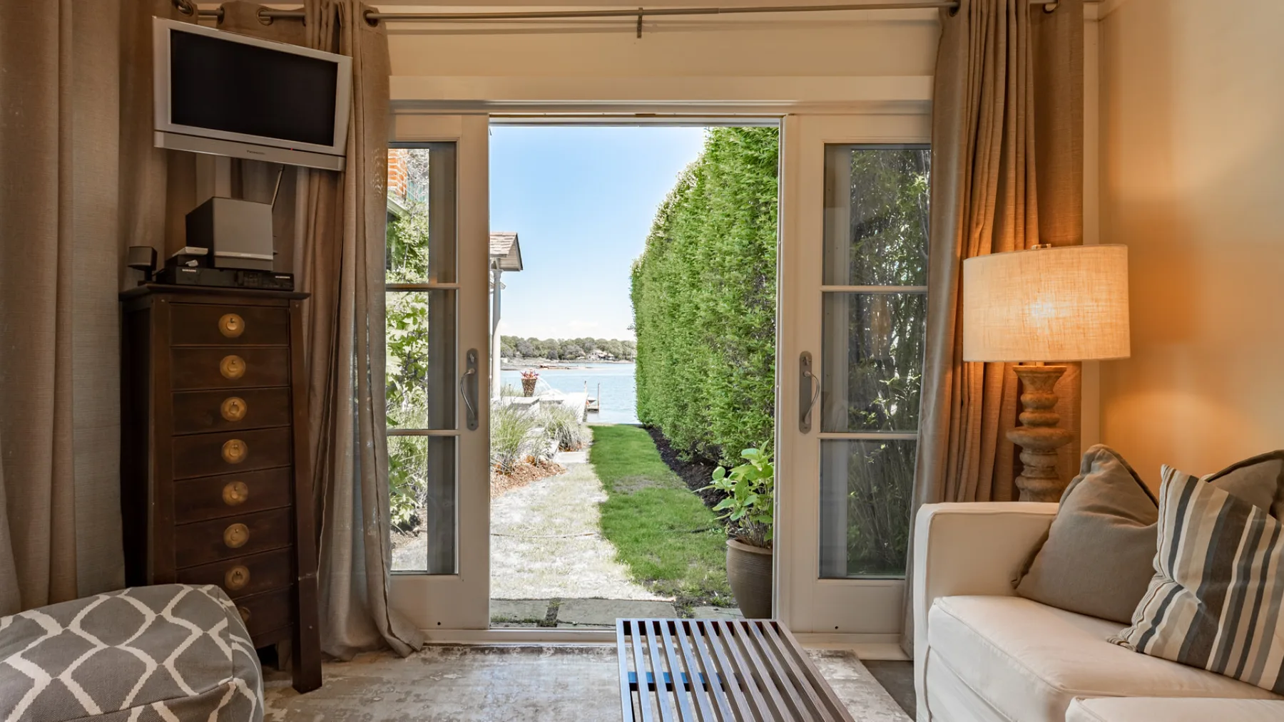 A living room opens through glass doors onto a garden path and water view.