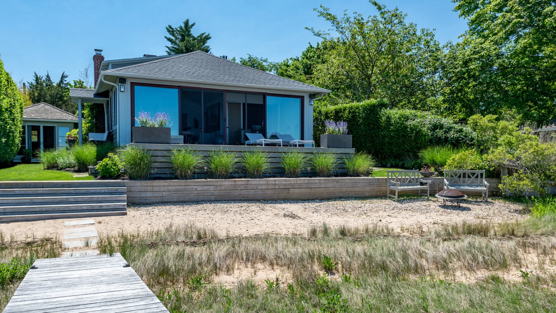 A modern house with large windows overlooks a sandy shoreline and landscaped garden.