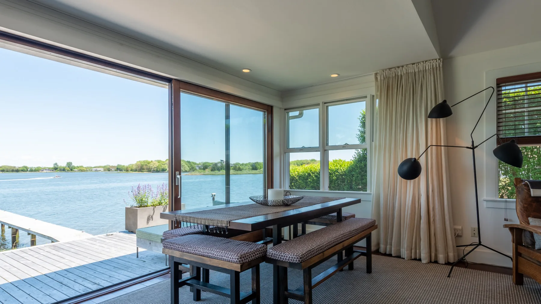 A dining area with a table and benches beside large windows overlooking the water.