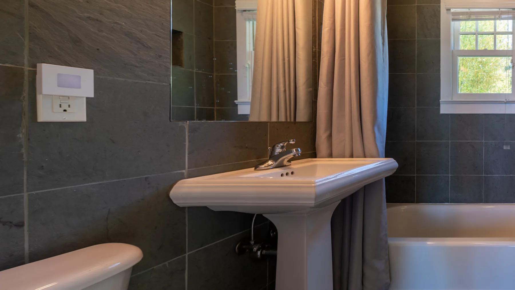A bathroom with a pedestal sink, a curtained window, and a bathtub.