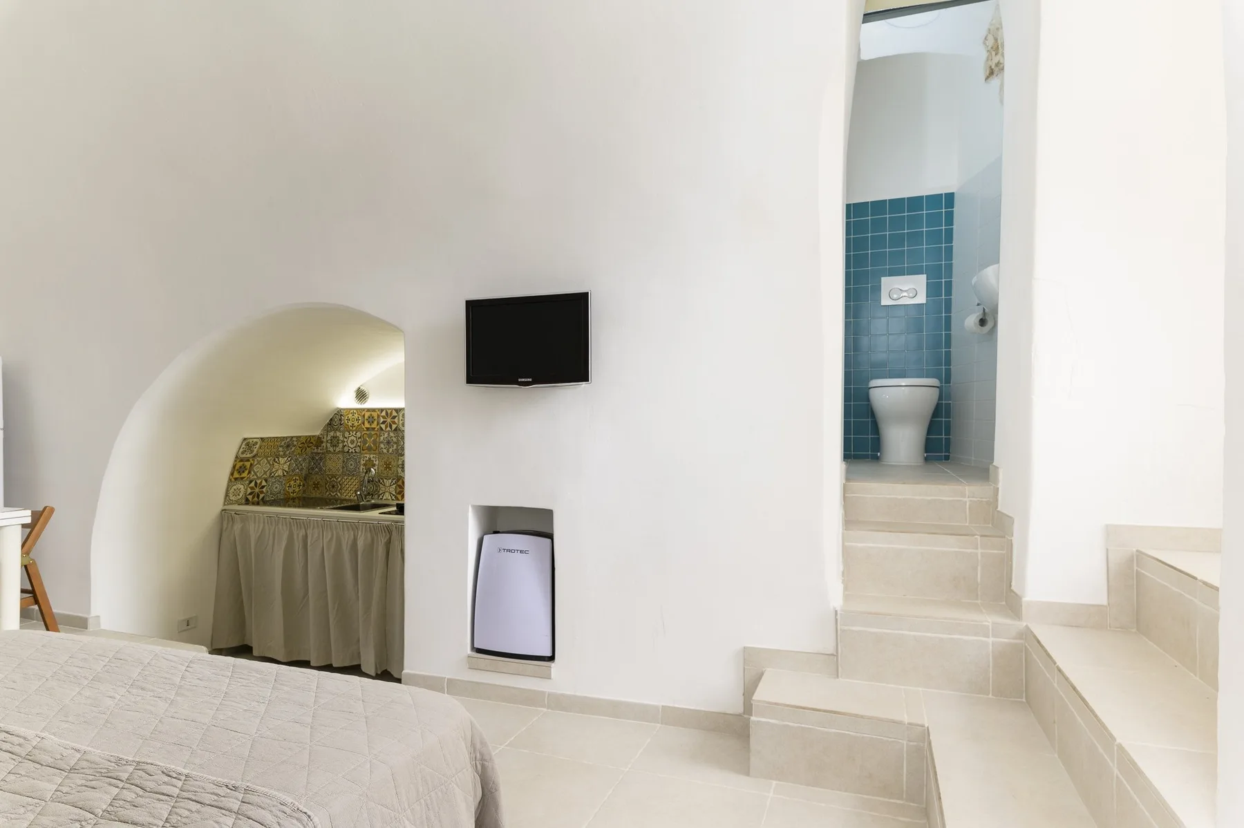 Un dormitorio con cama, televisor en la pared y vista abierta a un baño alicatado.