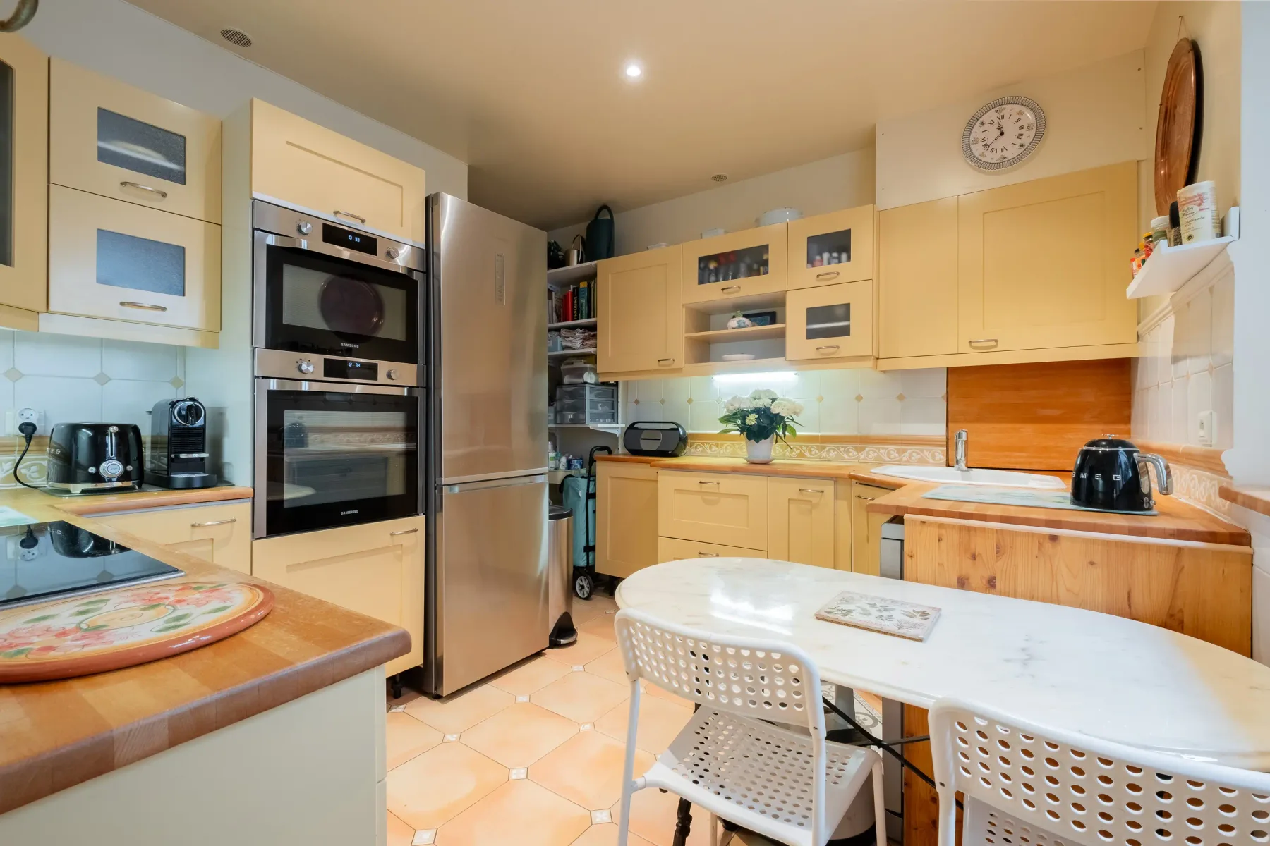 Una cocina con armarios de madera clara, hornos empotrados y una mesa blanca.