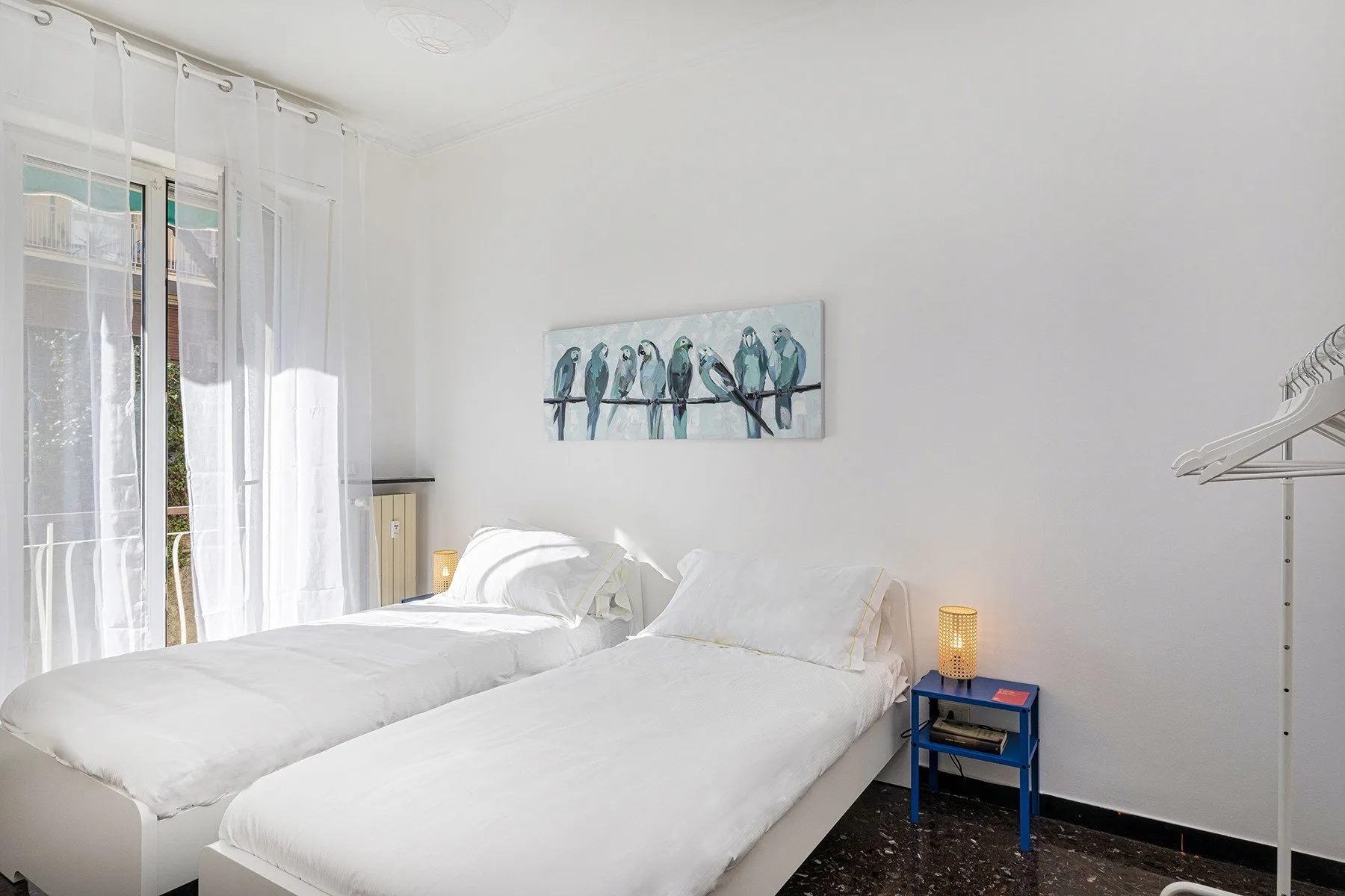 Un dormitorio luminoso con dos camas junto a ventanas altas con cortinas translúcidas.