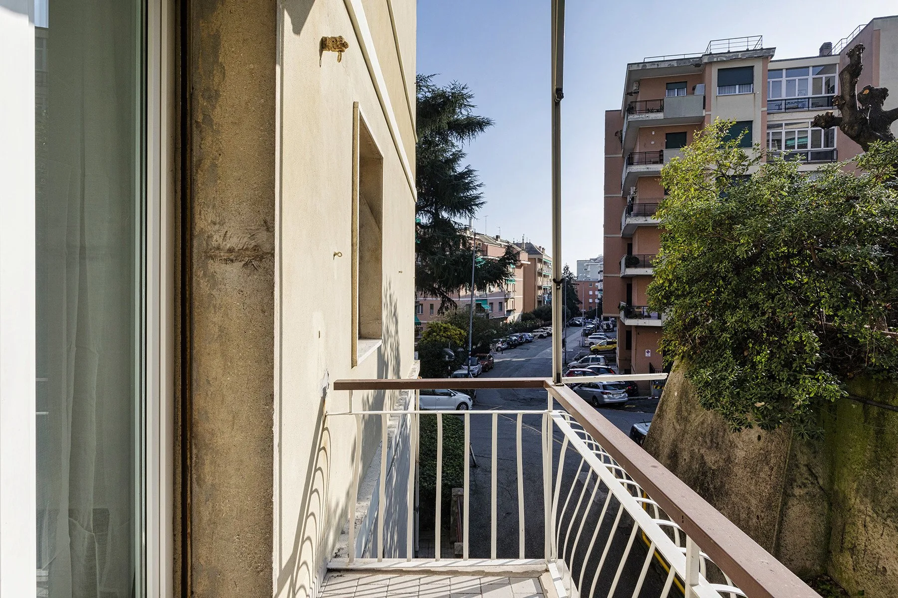 Un pequeño balcón da a una calle residencial con edificios de apartamentos y árboles.