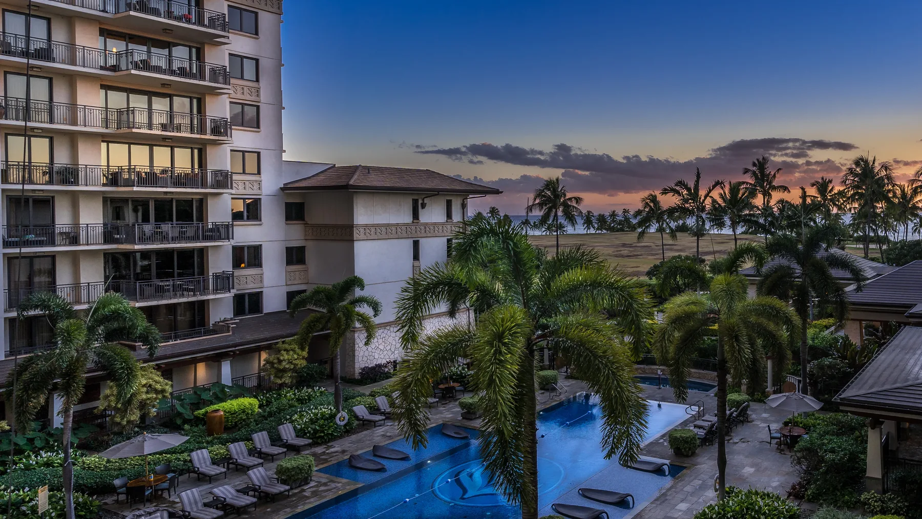 Una piscina rodeada de palmeras está junto a un edificio de varios pisos al atardecer.