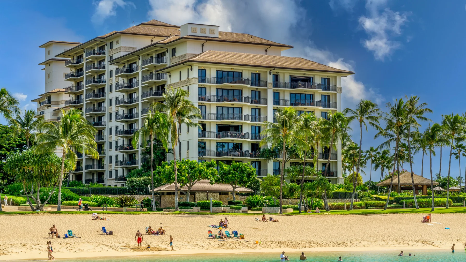 Un edificio junto a la playa se alza detrás de una playa de arena bordeada de palmeras.