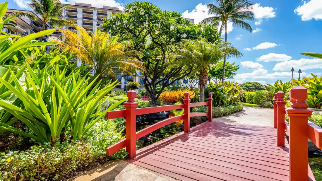 Un puente peatonal rojo cruza un jardín tropical ajardinado.