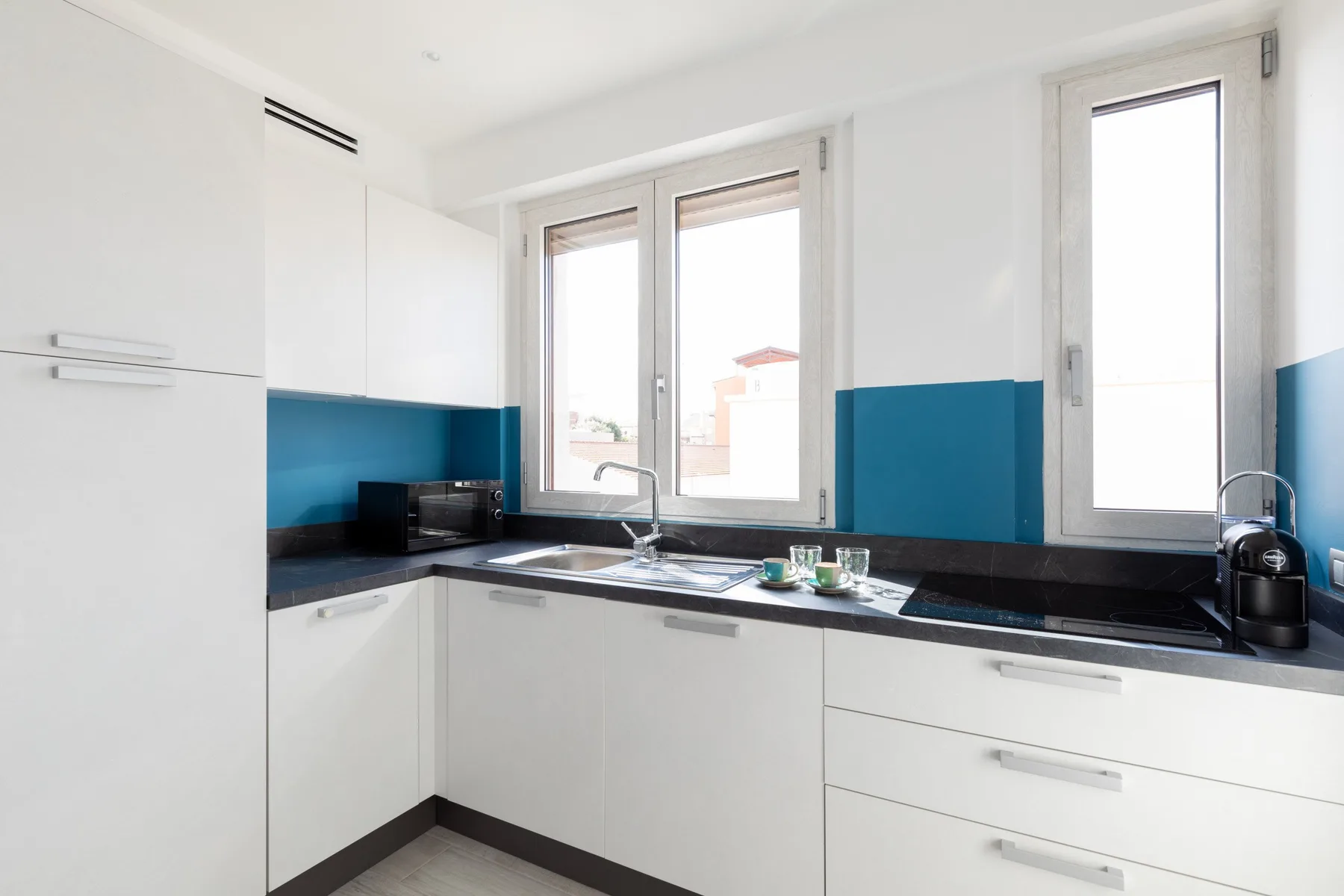 Una cocina luminosa con armarios blancos, salpicadero azul y un fregadero bajo las ventanas.