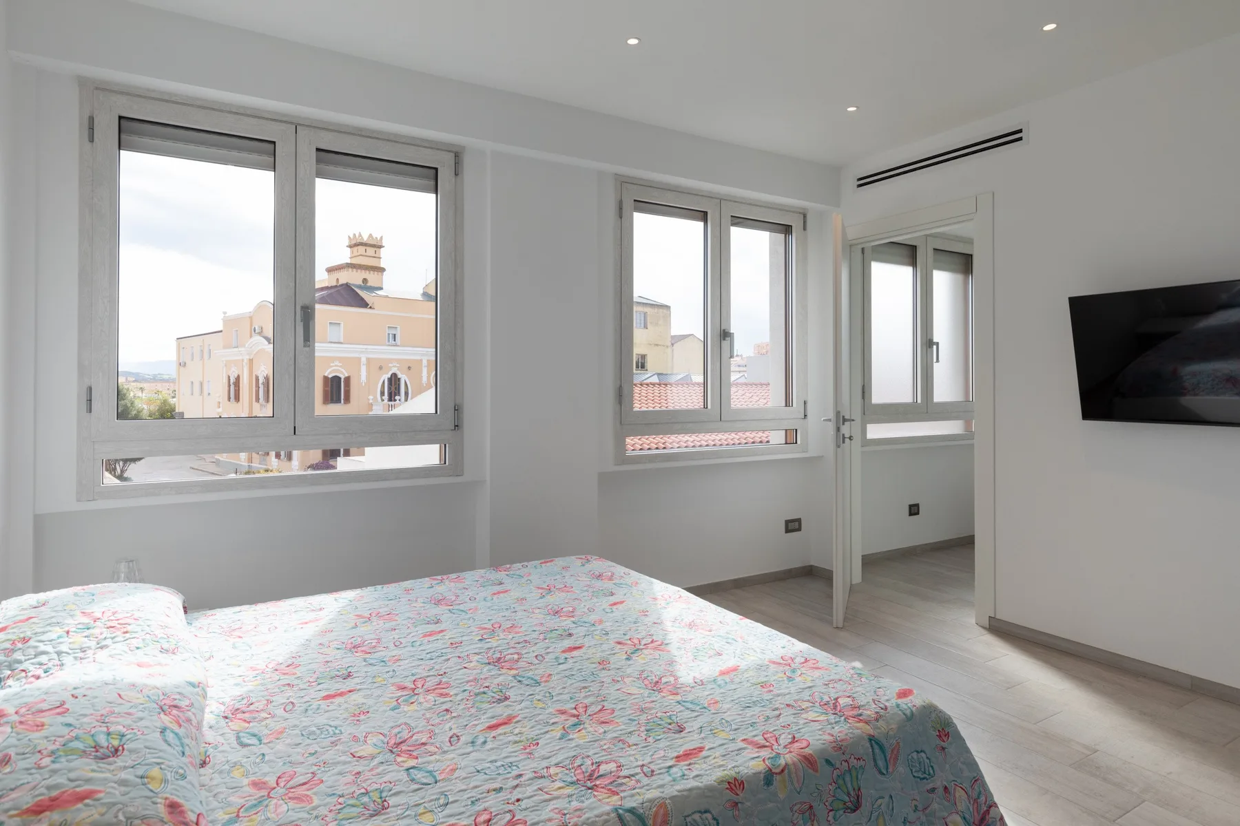 Un dormitorio luminoso con una colcha estampada, ventanas grandes y un televisor en la pared.