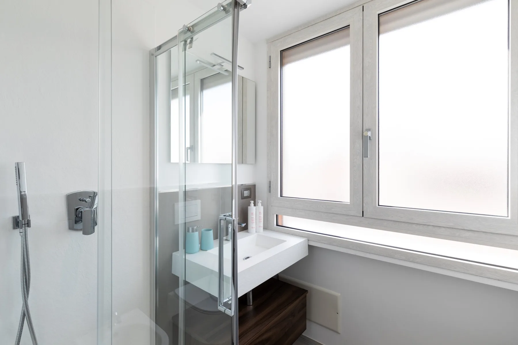 Un baño luminoso con ducha de vidrio, lavabo y una ventana grande.