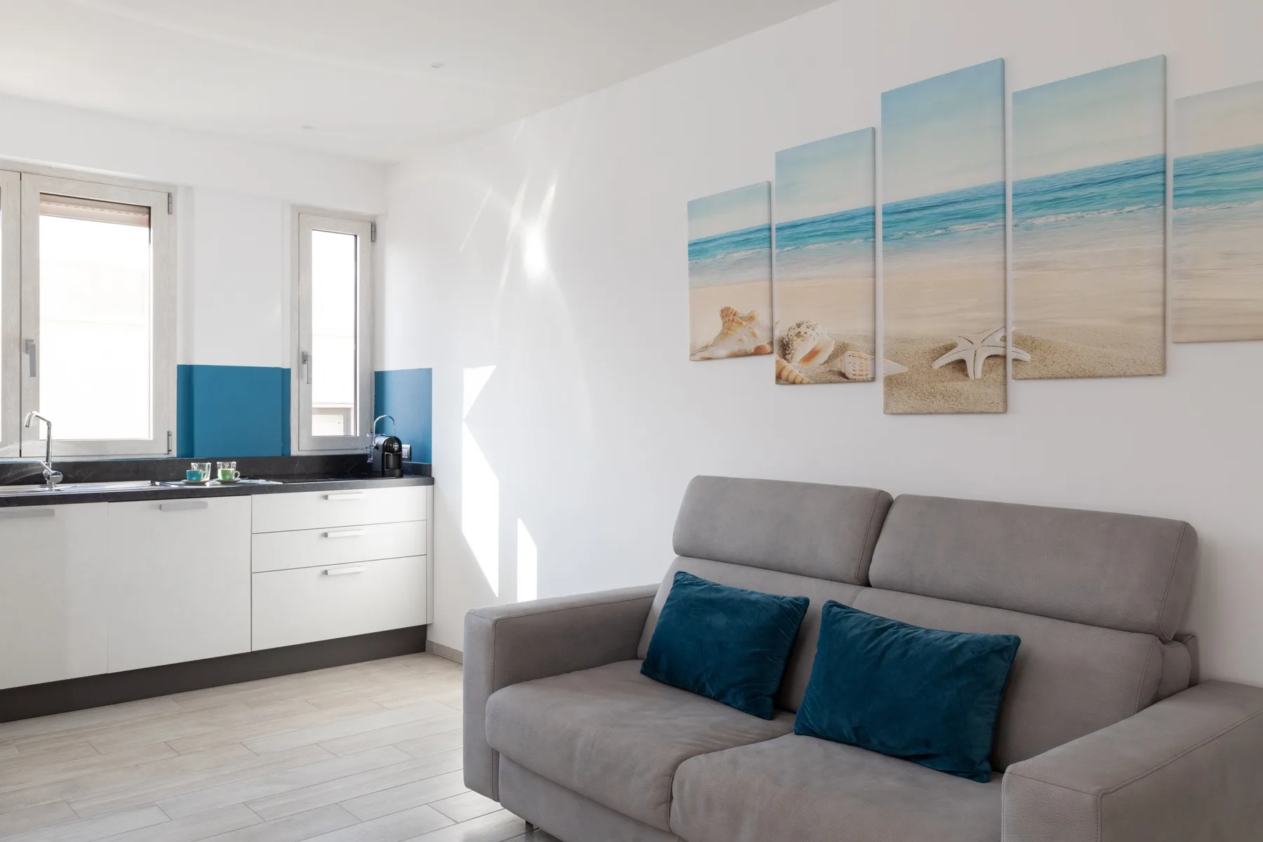 Una sala luminosa con un sofá gris y una pequeña cocina.