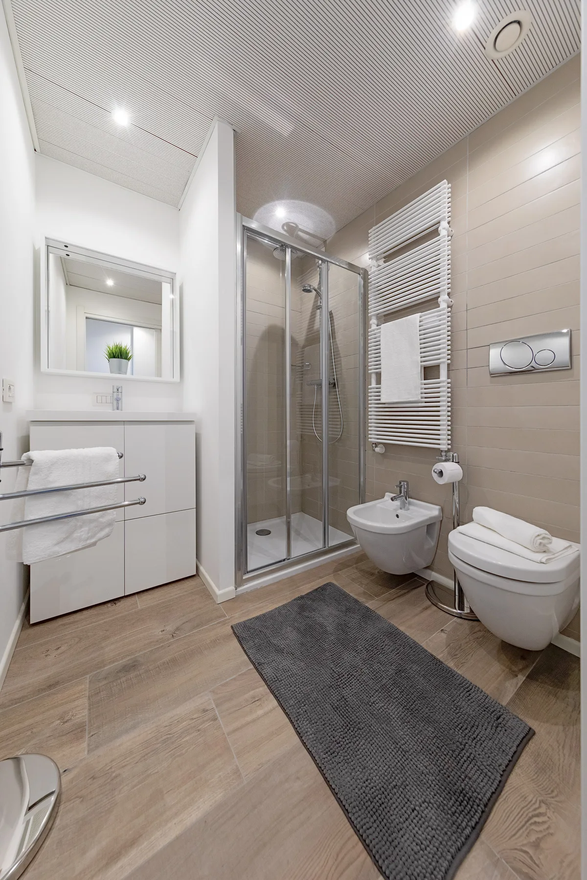 Un baño moderno con ducha de vidrio, lavabo suspendido, inodoro y alfombra gris.