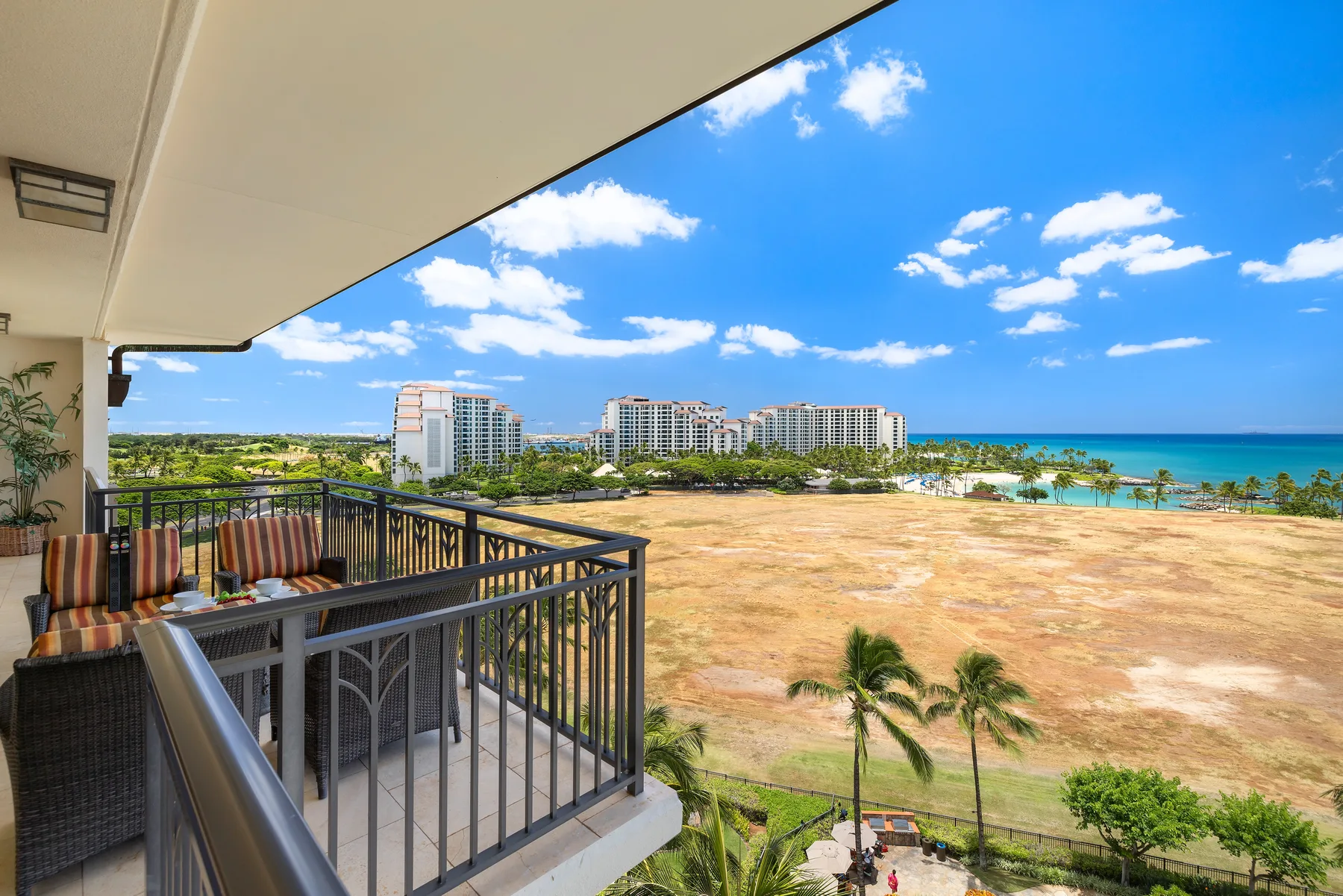 A balcony with outdoor seating overlooks an open field, palm trees, and the ocean.