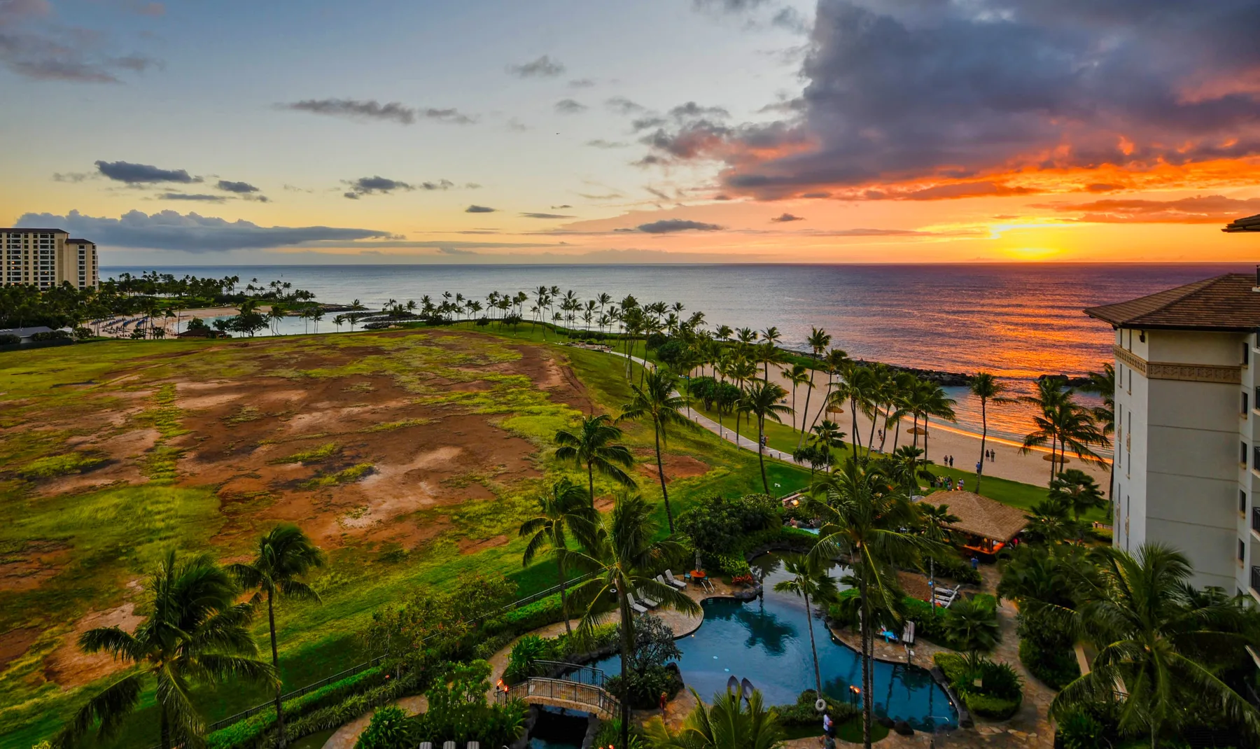 A coastal resort landscape at sunset, with palm trees and a pool beside the ocean.