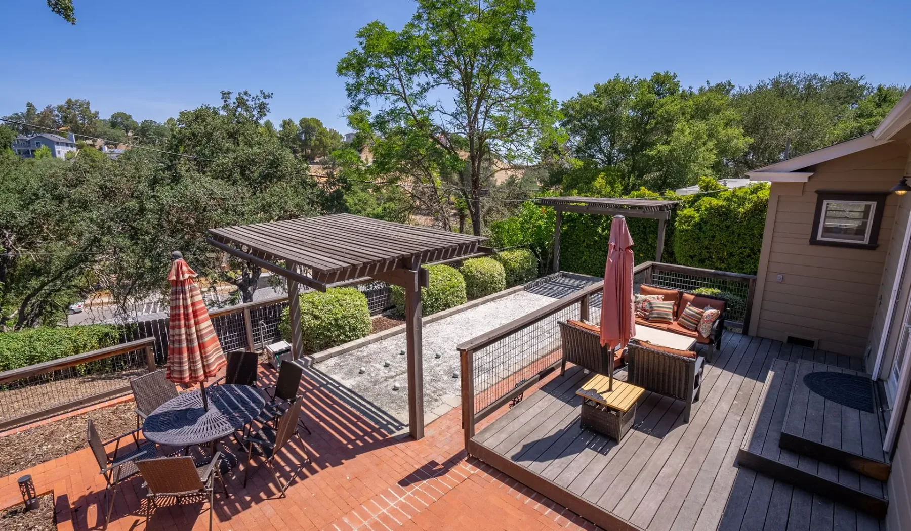 Una terraza exterior tiene asientos bajo una pérgola, rodeada de árboles.
