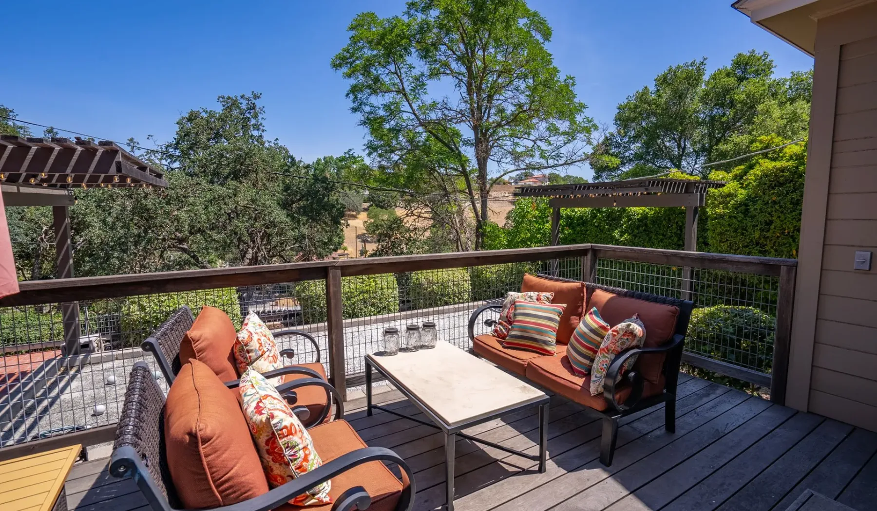 Una terraza exterior tiene asientos acolchados alrededor de una mesa baja, con árboles más allá de la barandilla.