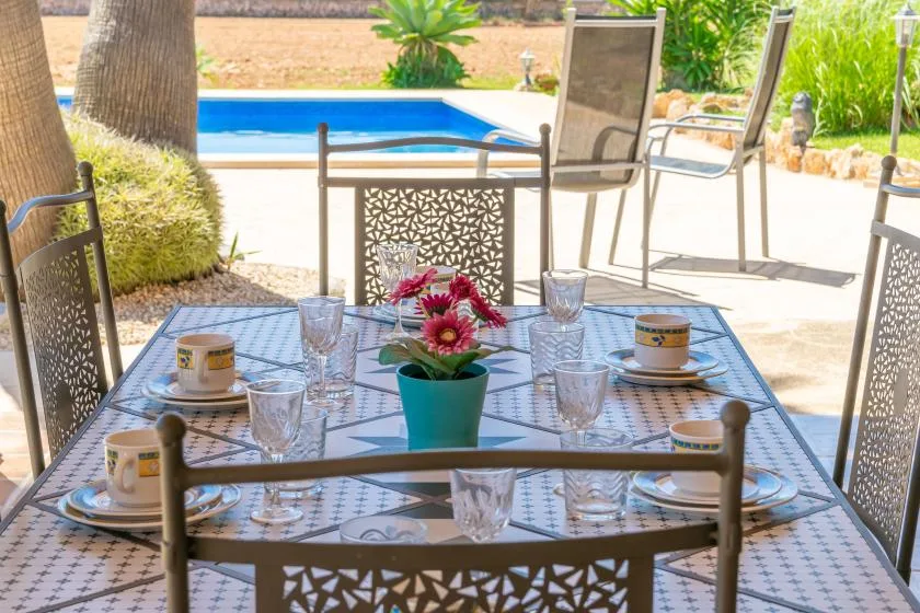 Una mesa de comedor en el patio está puesta con tazas y flores junto a una piscina.