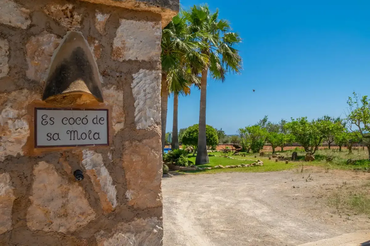 Un letrero de piedra está junto a un jardín soleado con palmeras y un camino de grava.