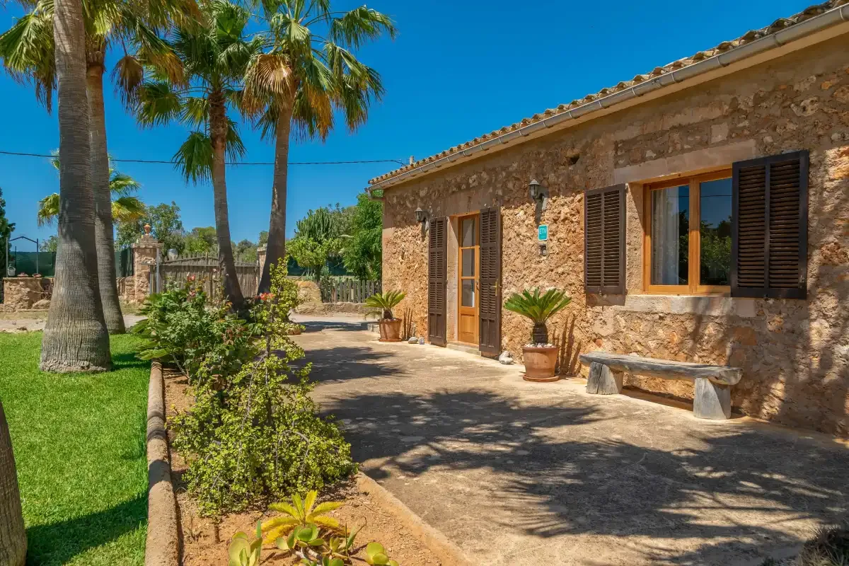 Exterior de piedra junto a un camino pavimentado, con palmeras y plantas de jardín.