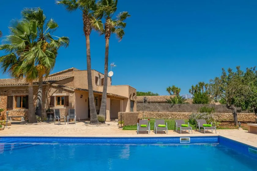 Una piscina frente a una casa baja, con palmeras y asientos al aire libre.