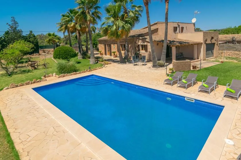 Una piscina al aire libre junto a una casa de color tostado, con palmeras y asientos de descanso.