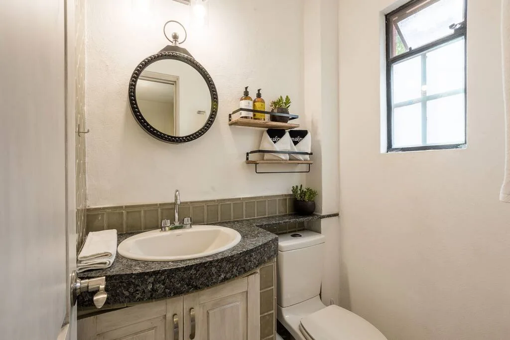 Bathroom with a round mirror, sink, and open shelves beside a window