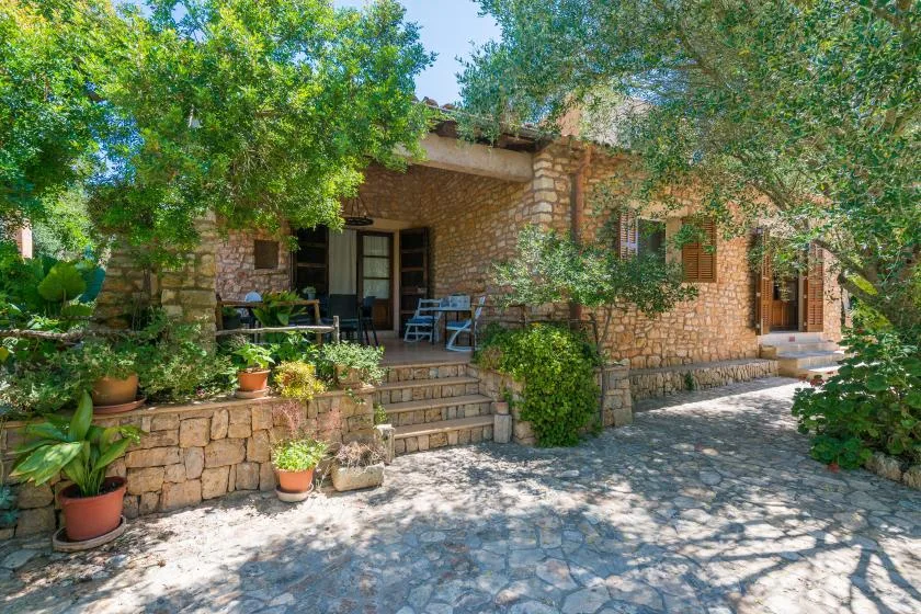 A stone house opens onto a shaded patio with outdoor seating and potted plants.
