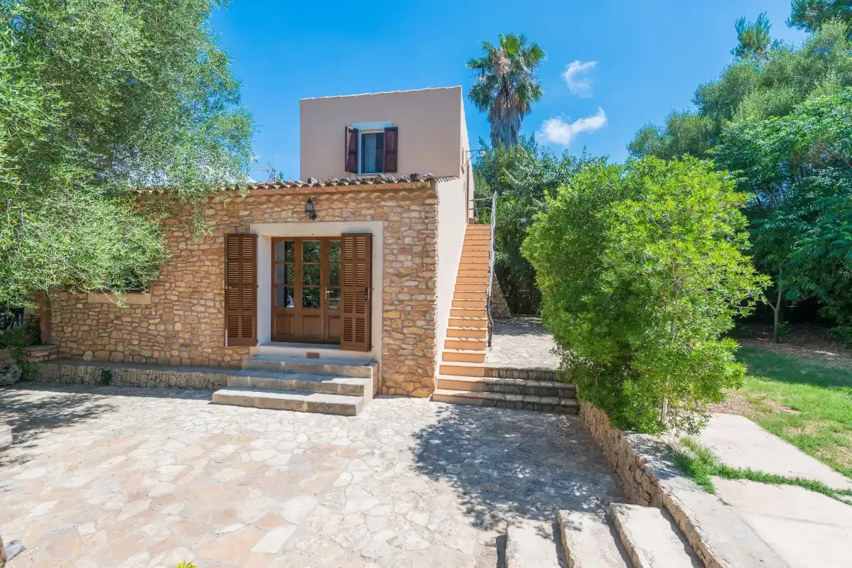 A stone house stands beside an outdoor staircase in a sunny courtyard.
