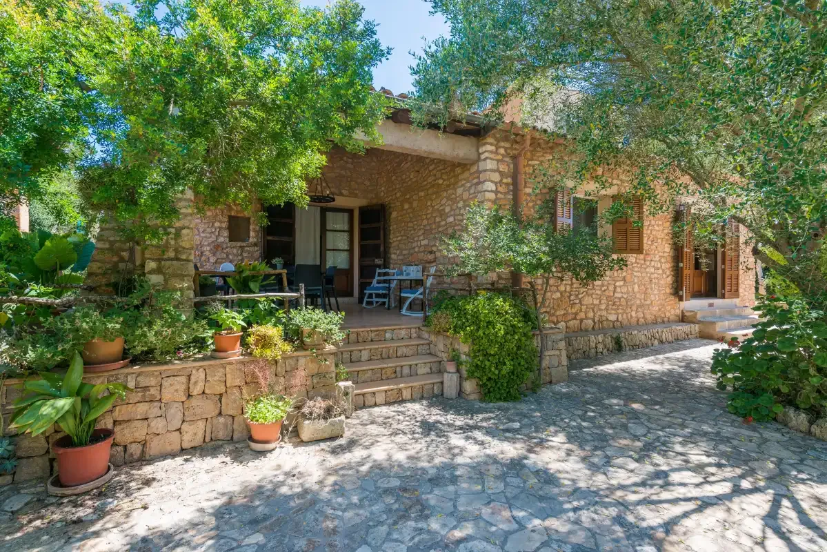 A stone house opens onto a shaded patio with outdoor seating and potted plants.