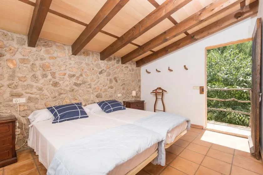 A bedroom with two beds, exposed wooden ceiling beams, and an open doorway to greenery.