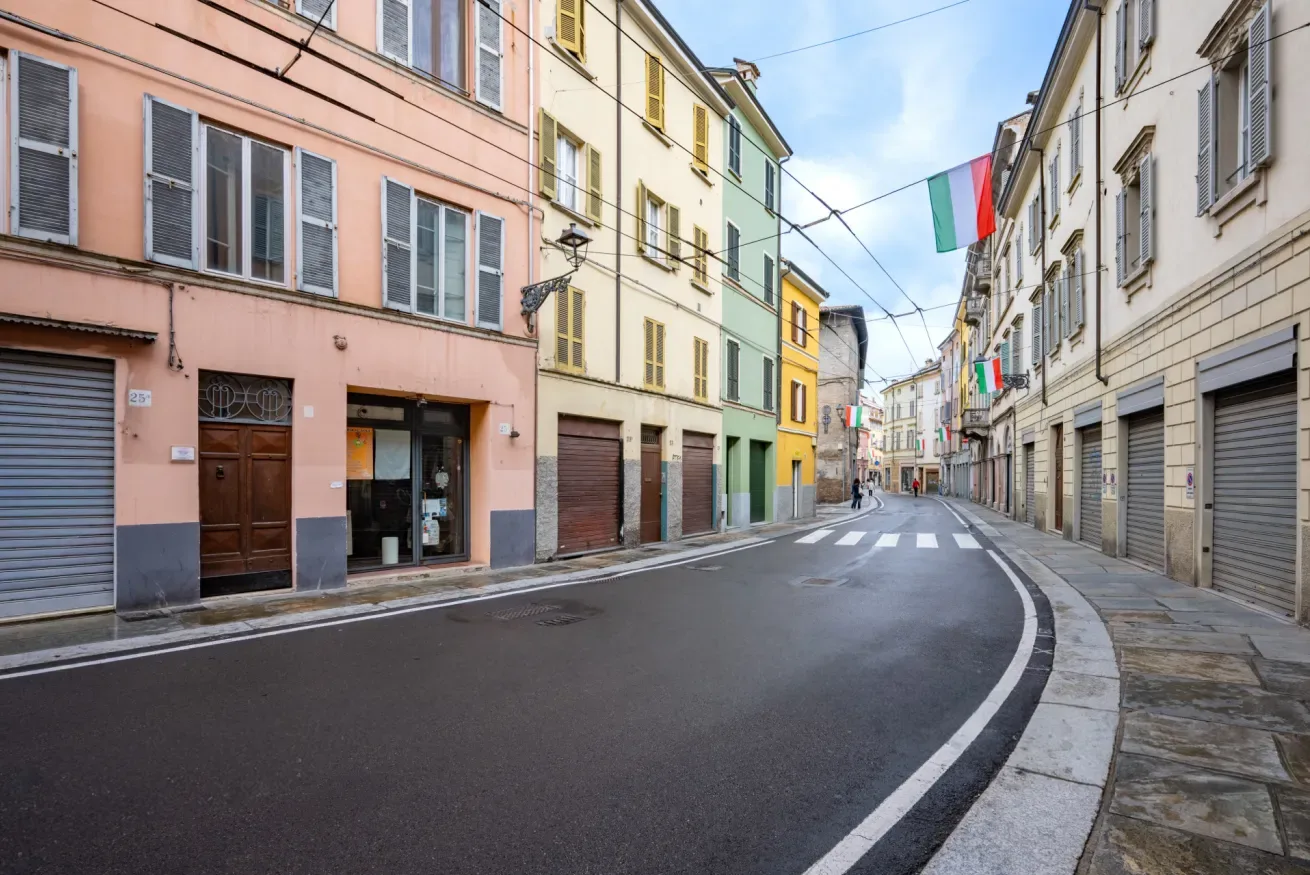 Una calle curva pasa entre filas de edificios de varias plantas, con banderas italianas sobre ella.