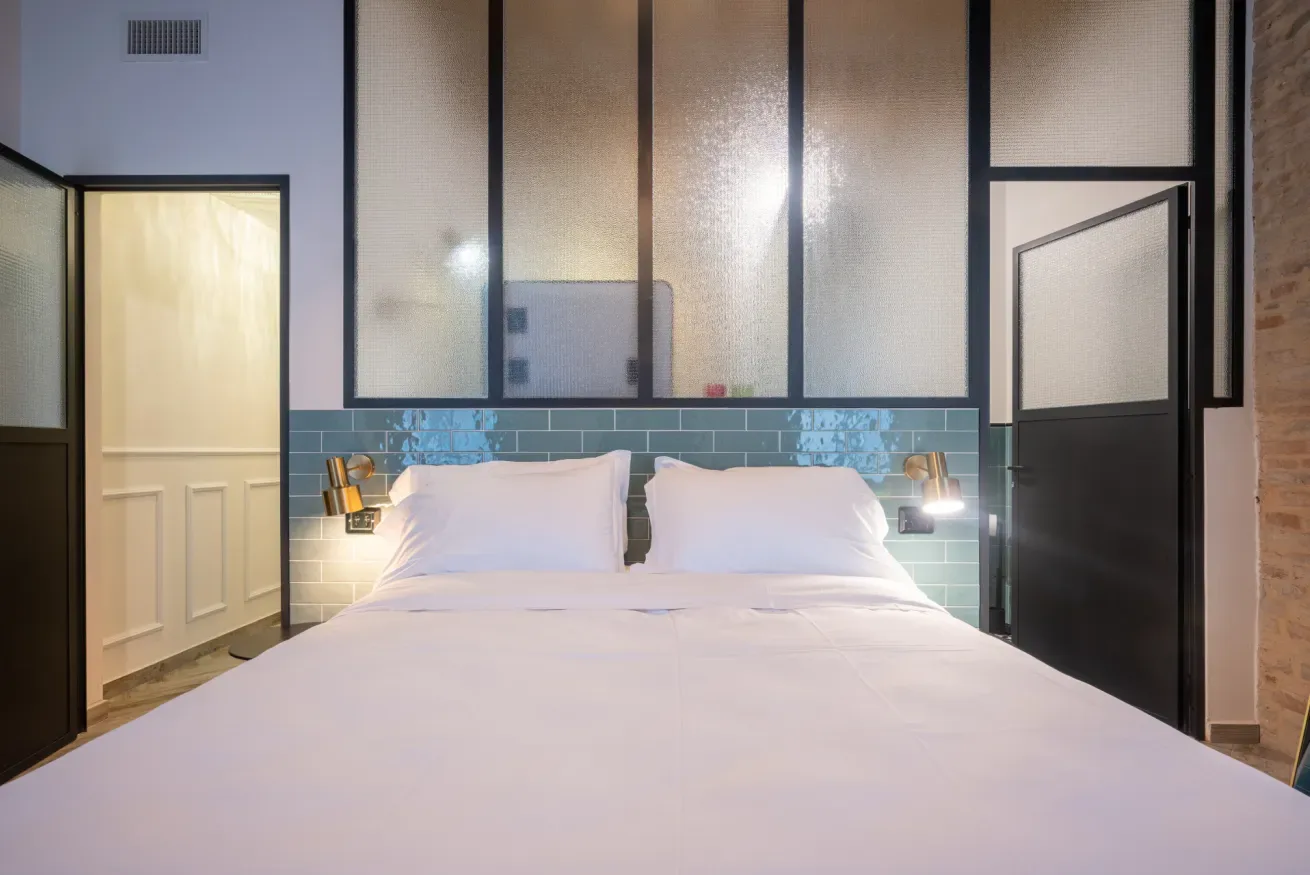 Un dormitorio con una cama vestida con sábanas blancas y luces de lectura en la pared.