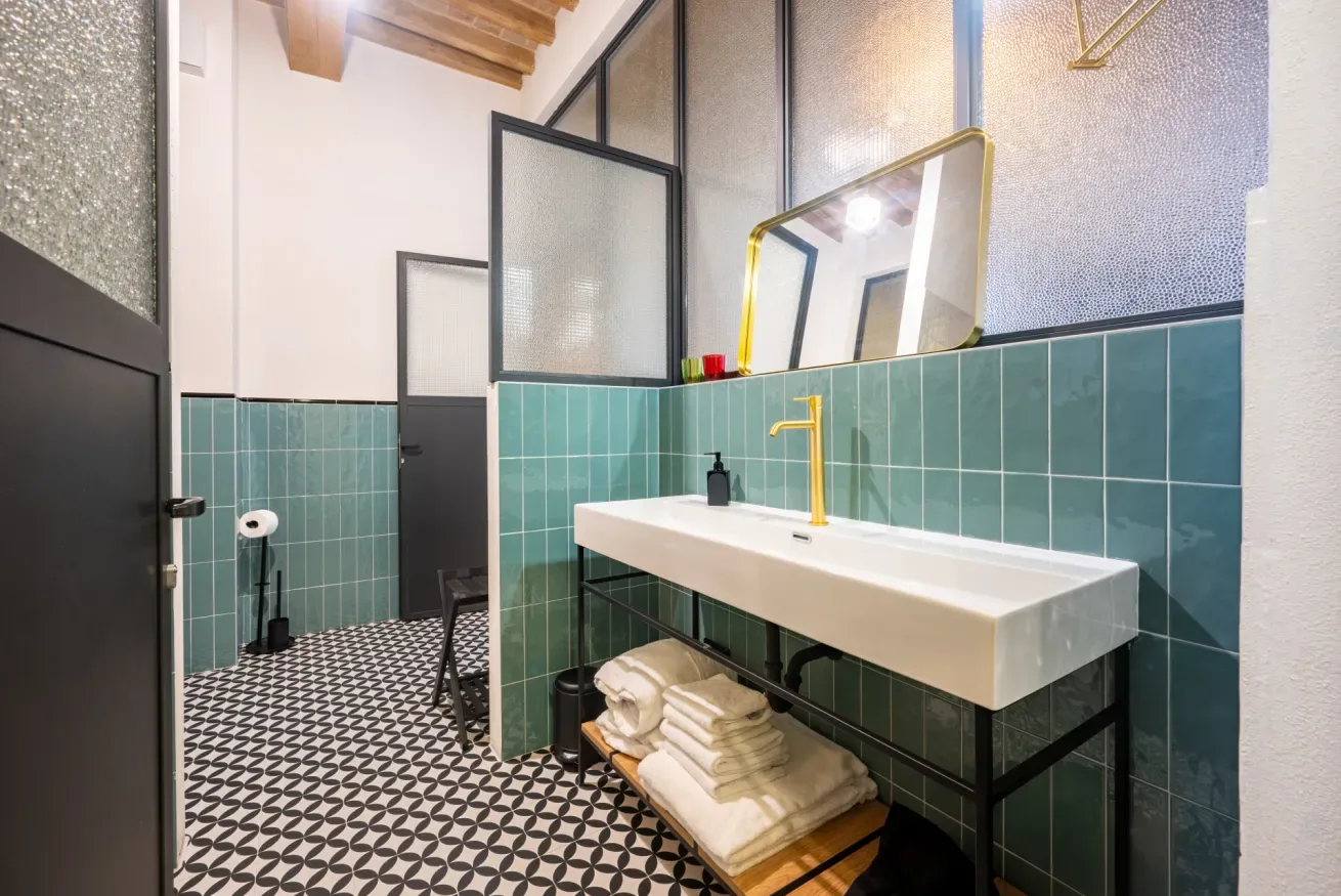 Un baño alicatado con un lavabo blanco largo, un grifo dorado y suelo estampado.