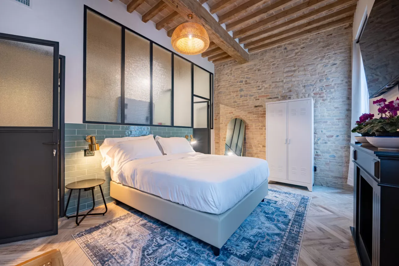 Un dormitorio con una cama bien tendida, pared de ladrillo visto y alfombra estampada.